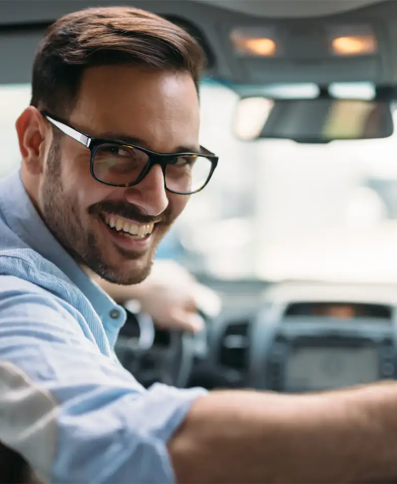 Motorista homem jovem simpático usando óculos, em seu carro sorrindo, virado para trás com o braço direito sobre o banco do passageiro, indicando extrema satisfação. Página: Recurso de Multa de Trânsito - Resolv Multas